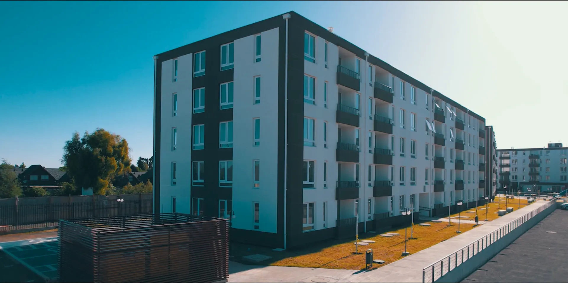 Conjunto habitacional de departamentos modernos de cinco pisos con balcones, rodeado de áreas verdes, veredas y estacionamientos, bajo un cielo despejado.